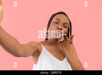 Young black woman taking selfie with cellphone, sending air kiss, filming content for blog on pink background Stock Photo