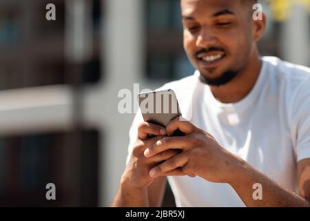 Closeup shot of african american man using cellphone, texting or surfing internet, sitting outdoors, focus on device Stock Photo