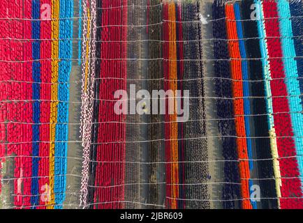 June 8, 2022, Narayanganj, Dhaka, Bangladesh: Workers hang thousands of meters of different colored fabrics to dry so they are able to be made into t-shirts and vests. Iron wires are used between bamboo frameworks to create giant washing lines for the final part of the dying process as the fabrics are dried in the sun in Narayanganj, Bangladesh. Bright strands of blue, pink, orange, red, purple, yellow, magenta and green-dyed cloths hang above the grassy field in a dazzling network of interlocking colors. After five to six hours in the sun, the fabric is used to make dresses.This is the final Stock Photo