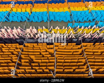 June 8, 2022, Narayanganj, Dhaka, Bangladesh: Workers hang thousands of meters of different colored fabrics to dry so they are able to be made into t-shirts and vests. Iron wires are used between bamboo frameworks to create giant washing lines for the final part of the dying process as the fabrics are dried in the sun in Narayanganj, Bangladesh. Bright strands of blue, pink, orange, red, purple, yellow, magenta and green-dyed cloths hang above the grassy field in a dazzling network of interlocking colors. After five to six hours in the sun, the fabric is used to make dresses.This is the final Stock Photo