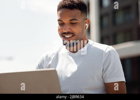 Handsome black man with wireless earphones working on laptop, freelancer sitting in urban city area Stock Photo