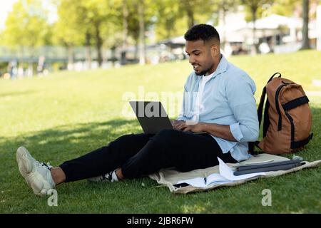 Black male freelancer using laptop computer outdoors, sitting on green grass in park and working remotely Stock Photo
