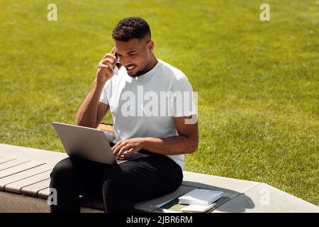 Positive african american man sitting outdoors in city, talking on cellphone while using laptop, working outside Stock Photo