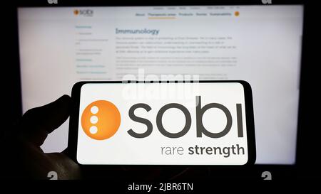 Person holding smartphone with logo of company Swedish Orphan Biovitrum AB (Sobi) on screen in front of website. Focus on phone display. Stock Photo