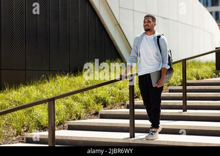 Happy african american man walking in urban city park with laptop and backpack, going down the stairs, copy space Stock Photo