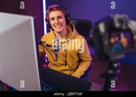 Happy young streamer pro gamer in headphones enjoying shooting vlog, smiling on camera, using microphone. Man pro gamer influencer streaming while playing computer video games. Esport and cyber sport Stock Photo
