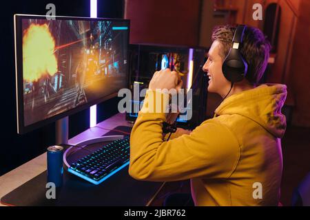 Side view of young caucasian pro gamer celebrating his victory in online video shooter game tournament. Exited cyber sportsman playing shooters on powerful pc at home at night, wearing headphones Stock Photo