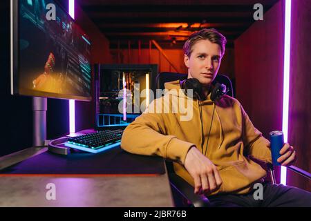 Portrait of young serious handsome pro gamer sitting at desk with gaming equipment, holding energy drink, using powerful PC for playing online video shooter games. Neon Colored Room, e-Sport concept  Stock Photo