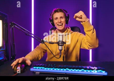 Handsome guy is screaming and keeps his hand up because of win in online tournament. Competitive gamer playing online video game at home, streaming playthrough video. Neon color. Cyber sport concept  Stock Photo