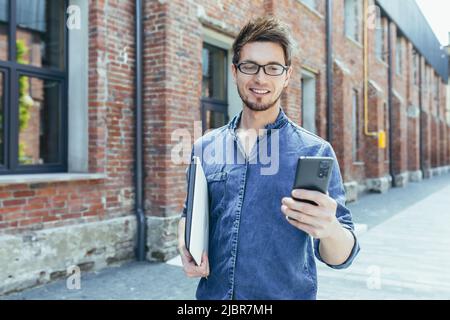 Portrait of young male student, outside campus holding cellphone, smiling and walking Stock Photo