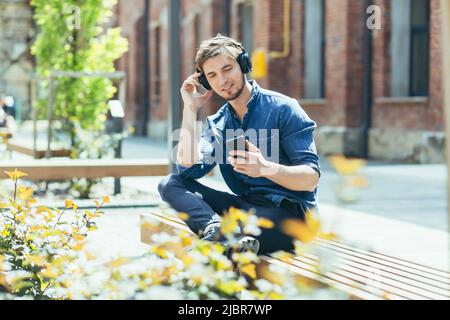 Young man sitting on a bench outside the office listening to music enjoys a mobile application for a smartphone, with large headphones Stock Photo