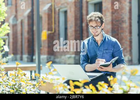 Portrait of a young teacher outside the university campus, sitting on a bench with a laptop and smiling Stock Photo