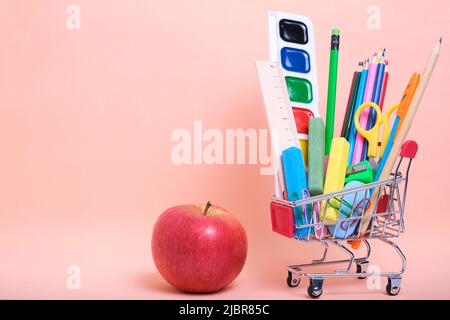 Shopping cart full school supplies and office supplies and red apple pink background Stock Photo