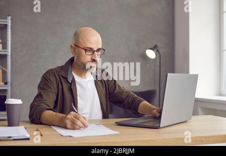 Man sitting at table in home office, working on laptop and writing down information Stock Photo