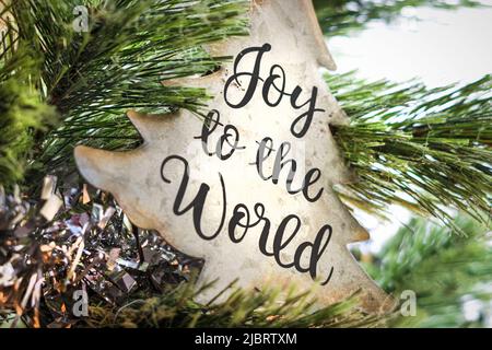 A Christmas tree shaped tin ornament with the words Joy to the World hanging in a lighted Christmas tree with tinsel Stock Photo