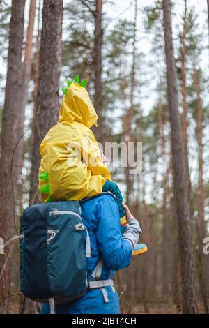 Mom and child walking in the forest after rain in raincoats together, the child is sitting on the shoulders of mom, back view Stock Photo