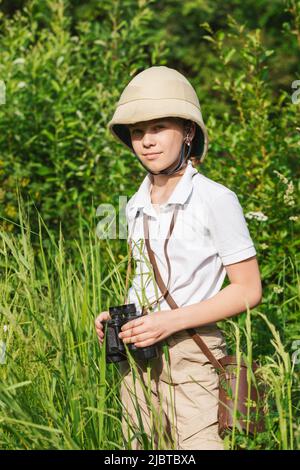 Preteen girl wearing pith helmet stands in the grass holding binoculars observing summer nature. Discovery and adventures concept Stock Photo