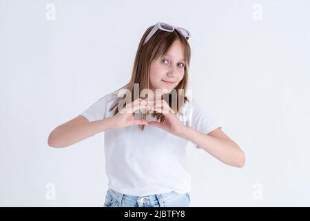 Stylish teen girl with long blond hair and blue eyes in a white t-shirt and blue jeans shows a heart shape with her fingers and smiles on a white back Stock Photo