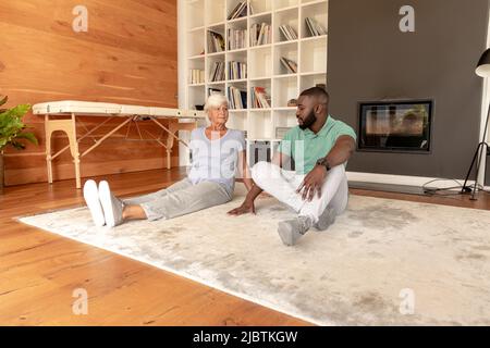 African american male physiotherapist and caucasian senior woman sitting on the floor at home Stock Photo