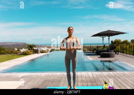 Caucasian young woman practicing yoga and meditating near the pool Stock Photo