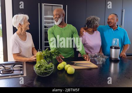 Happy multiracial seniors with apples, leaf vegetables and utensils on counter talking in kitchen Stock Photo
