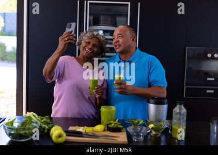 African american senior woman taking selfie with caucasian man while having smoothie in kitchen Stock Photo