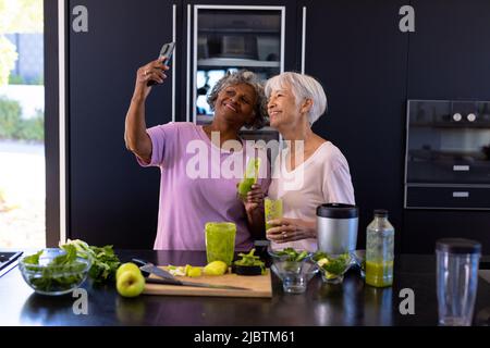 African american senior woman taking selfie with asian friend while holding smoothie in kitchen Stock Photo