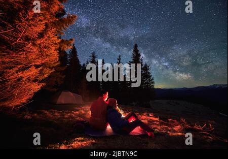 Couple in love sitting on mat, admiring fairytail starry sky with Milky way. Romantic cozy atmosphere in the fresh air against backdrop of tent, trees, mountain hills and millions of stars. Stock Photo