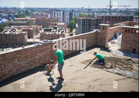 Man builder using troweling machine while screeding floor on the roof of residential building. Another man leveling surface with straight edge while screeding floor outdoors in new building. Stock Photo