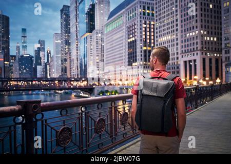 Rear view of man with backpack while walking on bridge and looking around. Illuminated city with skyscrapers at twilight. Stock Photo