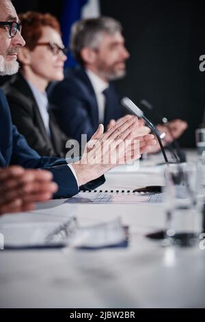 Senior buisnessman in eyeglasses and suit clapping hands together with his colleagues while they sitting at table at business conference Stock Photo