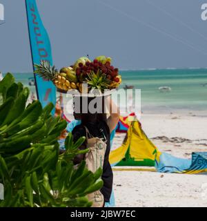 Paje, Zanzibar, Tanzania - January 202: African man with a tray full of exotic fruits on his head. Surfing beach in Paje in the background, Africa Stock Photo