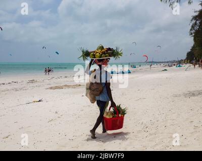 Paje, Zanzibar, Tanzania - January 202: African man with a tray full of exotic fruits on his head. Surfing beach in Paje in the background, Africa Stock Photo