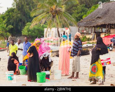 Kizimkazi, Zanzibar - February 2021: People in colorful African costumes wait for fishermen and fish at a small fish market on a sandy beach. Sunset t Stock Photo