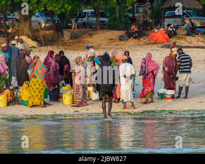 Kizimkazi, Zanzibar - February 2021: People in colorful African costumes wait for fishermen and fish at a small fish market on a sandy beach. Sunset t Stock Photo