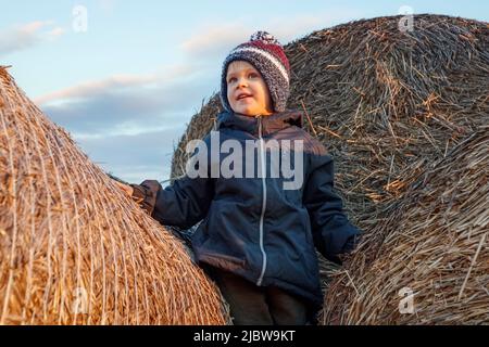 A smiling cute boy in a knitted hat stands on a golden stack of straw in the evening sun. The child's face is illuminated by the evening sun, he is cl Stock Photo