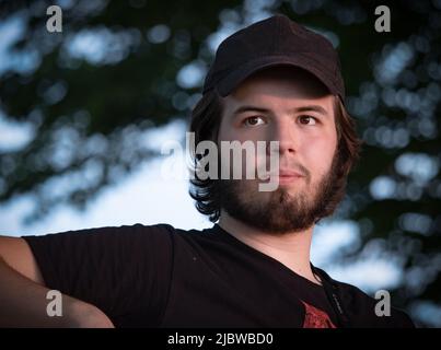 Young adult man with longer dark hair, dark eyes, dark beard,and moustache with a baseball cap sitting at the park contemplating in summer Stock Photo