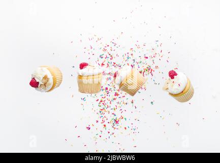 On a white background with multi-colored confetti, small muffins with cream and raspberries in a frozen flight. Creative composition. Holiday, anniver Stock Photo