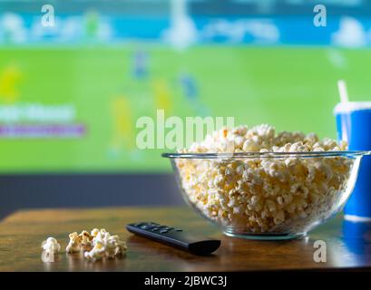 Popcorn in a glass bowl on the table and a TV remote control against the background of a large TV screen with a sports match. Cozy viewing of sports p Stock Photo