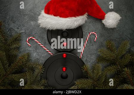 Dumbbells weight plates as snowman with red Santa Claus hat, candy canes hands, baubles as buttons. Gym fitness Christmas concept with tree branches. Stock Photo