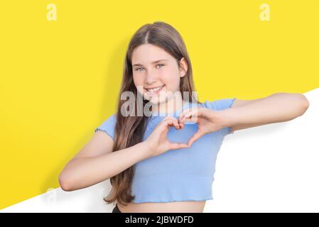 Young Caucasian girl with blue eyes smiling and forming a heart with her hands on white and yellow background. Gesture and expression of love Stock Photo