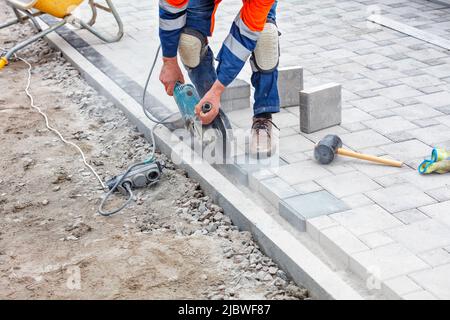 A construction worker using an electric grinder and a diamond cutting disc cuts paving slabs while paving a sidewalk in the daytime. Copy space. Stock Photo