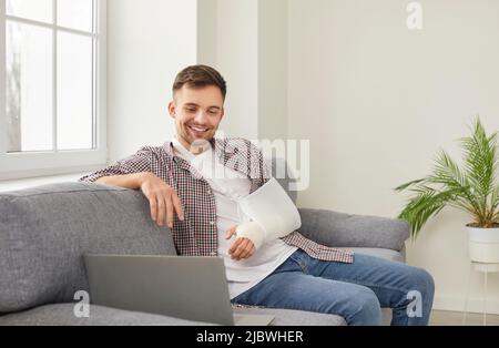 Happy young man with broken arm sitting on sofa at home and watching movie on laptop Stock Photo
