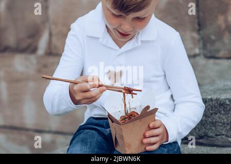 The child eating on the go soy sauce pan-fried noodles chopsticks. Dirty soy sauce stains on a white t-shirt. Takeaway food. outdoors Stock Photo