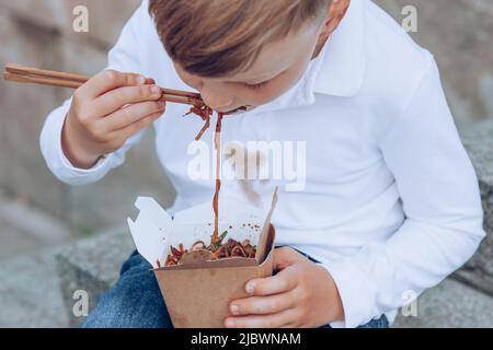 The child eating on the go soy sauce pan-fried noodles chopsticks. Dirty soy sauce stains on a white t-shirt. Takeaway food. outdoors Stock Photo