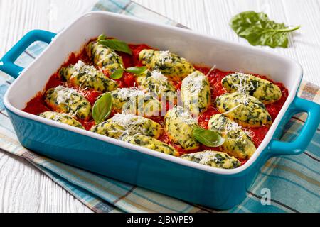 Malfatti, Italian spinach ricotta dumplings  in tomato sauce with herbs and grated parmesan cheese in ceramic baking dish on white table Stock Photo