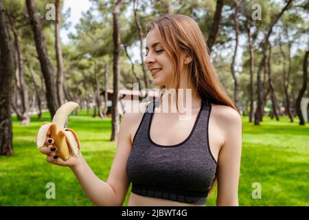 Portrait of young brunette woman wearing sports bra standing on city park, outdoors eating healthy banana for strength and energy. Outdoor sports and Stock Photo
