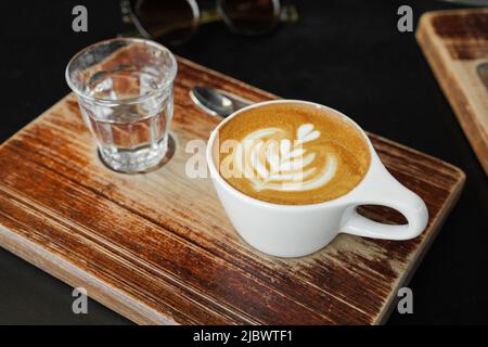 Cup of cappuccino with latte art in cozy cafe. Coffee and glass of water on wooden board Stock Photo