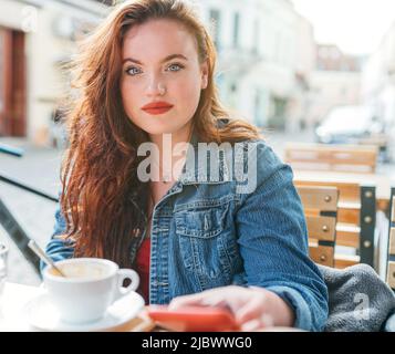 Portrait of sad red curled long hair caucasian woman sitting on a cozy cafe outdoor terrace on the street and looking at the camera. Natural beauty co Stock Photo