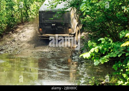 Back view of green russian off-road utility vehicle UAZ Hunter going up dirty road crossing river in forest among trees. Stock Photo